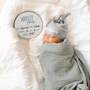 May include: A white round wooden plaque with black lettering announcing the birth of a baby named Wrigley James. The plaque includes the date, time, weight in pounds and ounces, and length in inches. A baby wearing a gray knitted hat with the name Wrigley on it is sleeping on a white blanket.
