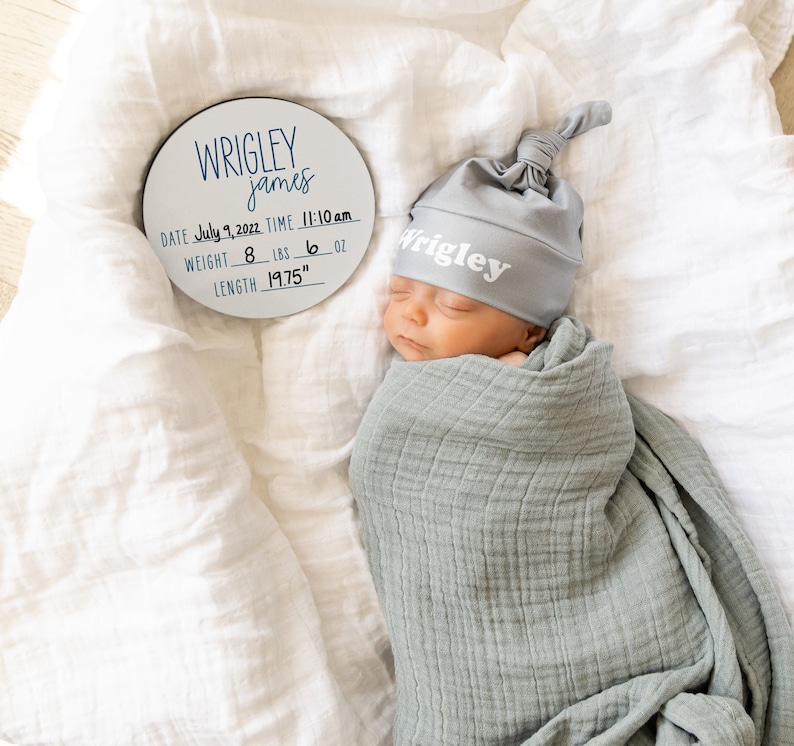 May include: A white round wooden plaque with black lettering announcing the birth of a baby named Wrigley James. The plaque includes the date, time, weight in pounds and ounces, and length in inches. A baby wearing a gray knitted hat with the name Wrigley on it is sleeping on a white blanket.