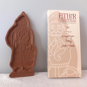 May include: A brown Father Christmas cookie mold with a Santa Claus design, holding a basket. The mold is next to a box with the text "Father Christmas" and "The 1990 Longaberger Pottery Cookie Mold".