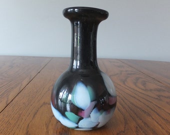 May include: A small black glass vase with a round body and a narrow neck. The vase is decorated with white, blue, green, and pink abstract shapes.