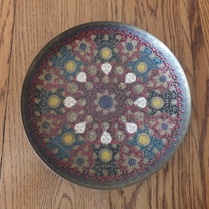 May include: A round, decorative brass plate with a red, gold, and white floral design. The plate has a raised, embossed pattern and a shiny finish.