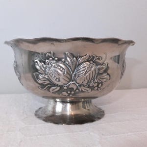 K&ouml;nnte beinhalten: Eine silberne Sch&uuml;ssel mit einem gewellten Rand und einem Fu&szlig;. Die Sch&uuml;ssel ist mit einem gepr&auml;gten Blumendesign verziert.