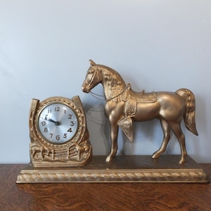 May include: A gold-colored horse-themed clock with a horse statue and a clock face. The clock face has black numbers and hands. The horse statue is saddled and stands on a rectangular base.