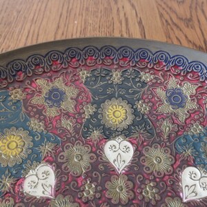 May include: A round brass tray with an intricate embossed design featuring floral patterns in shades of red, blue, green, gold, and white.