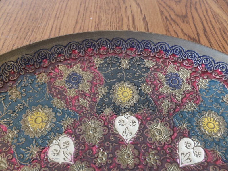 May include: A round brass tray with an intricate embossed design featuring floral patterns in shades of red, blue, green, gold, and white.