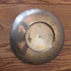 May include: A round, brass-coloured plate with a slightly raised centre. The plate has a smooth, shiny surface and a slightly worn appearance.