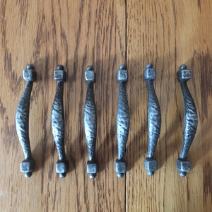 May include: Set of six rustic metal cabinet pulls. Each pull has a textured, hammered appearance with a curved handle and square ends. The pulls are silver-toned and arranged on a wooden surface.