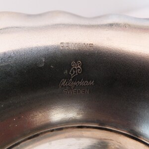 K&ouml;nnte beinhalten: Nahaufnahme einer silbernen Metallschale mit dem Text "PRMANS" und "GilleJohan SWEDEN", der auf den Boden graviert ist.