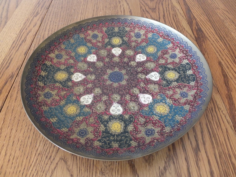 May include: A round brass tray with an intricate design featuring a floral pattern in shades of red, blue, yellow, and white.