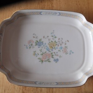 May include: A white ceramic serving dish with a gold rim and a floral design. The dish has a square shape with rounded corners.