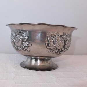 K&ouml;nnte beinhalten: Eine silberne Serviersch&uuml;ssel mit einem gepr&auml;gten Blumendesign. Die Sch&uuml;ssel hat einen gewellten Rand und einen Sockel.