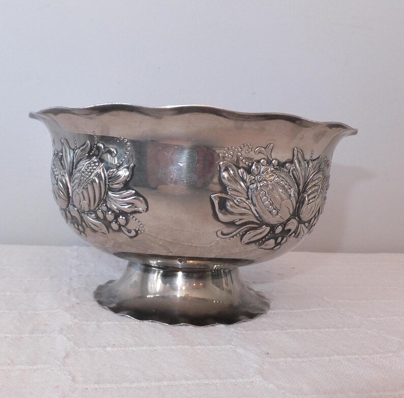 K&ouml;nnte beinhalten: Eine silberne Serviersch&uuml;ssel mit einem gepr&auml;gten Blumendesign. Die Sch&uuml;ssel hat einen gewellten Rand und einen Sockel.