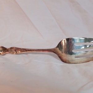 May include: A silver serving fork with an ornate handle. The fork has three tines and a slightly curved bowl.