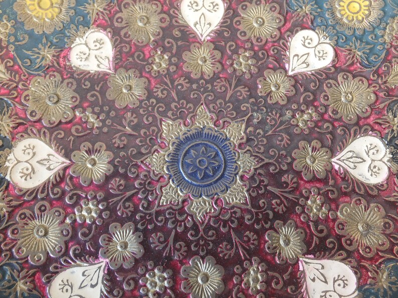 May include: A close-up of a decorative round tray with a red background and intricate gold and white floral designs. The centre of the tray features a blue and gold starburst pattern.