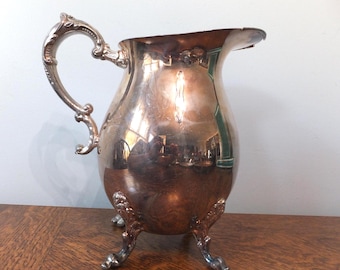 May include: A polished silver-toned metal pitcher featuring a curved handle with ornate detailing, a wide pouring spout, and three decorative feet. The reflective surface shows distorted surroundings, and the body has a rounded, bulbous shape.