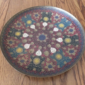 May include: A round brass tray featuring an intricate floral design in shades of red, gold, blue, and white. The tray has a raised rim and a decorative border.