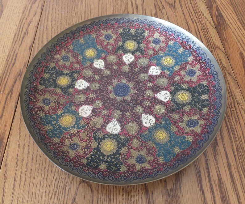 May include: A round brass tray featuring an intricate floral design in shades of red, gold, blue, and white. The tray has a raised rim and a decorative border.