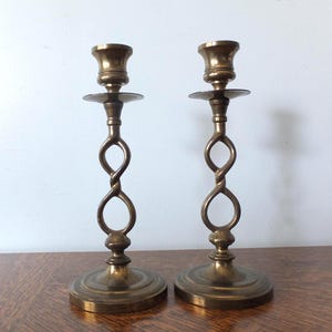 May include: Two antique brass candlesticks. Each candlestick has a round base, a twisted stem with an open loop design, and a cup-shaped top to hold a candle. The brass has a warm, aged patina.