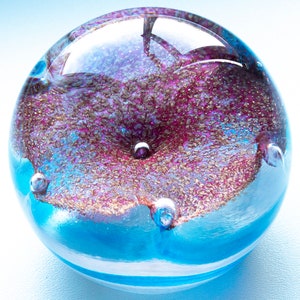 May include: A glass paperweight with a blue and pink swirl design and air bubbles. The paperweight is round and has a shiny finish.