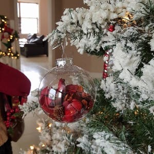 Peut inclure: Une boule de Noël en verre transparente remplie de pot-pourri rouge et brun. La boule est suspendue à un sapin de Noël artificiel enneigé.