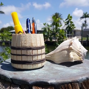 Op de afbeelding: Een houten tonvormige potloodhouder met een bruine touwrand, met een gele markeerstift, een blauwe pen, een zwarte pen en een rode pen. De houder staat op een houten oppervlak naast een grote witte schelp.