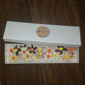 Op de afbeelding: Een witte doos met een logo dat "Augusta Dreams" en een halve maan zegt. De doos bevat een set van 12 kleine, kleurrijke, bloemvormige ornamenten gemaakt van schelpen.