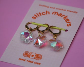 May include: Three iridescent crystal stitch markers with gold rings. The markers are shaped like flowers and are labeled "stitch markers" on a pink and white card with the text "Indie Yarn Club".