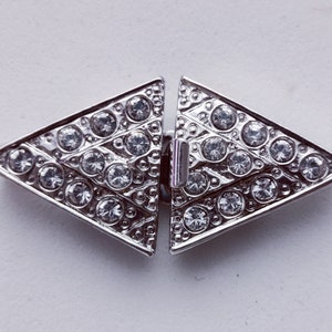 Könnte beinhalten: Ein silberfarbener, dreieckiger Verschluss mit geometrischem Design. Jedes Dreieck ist mit mehreren klaren, runden Strasssteinen verziert, die einen funkelnden Effekt erzeugen. Der Verschluss dient zum Verbinden von zwei Teilen.