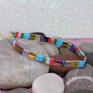 Könnte beinhalten: Ein farbenfrohes Perlenarmband mit rechteckigen Perlen in Gold, Blau, Rot und Grün. Das Armband ist flexibel und aufgereiht, ruhend auf einem Bett aus glatten Steinen.