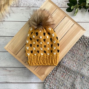 May include: A mustard yellow knit beanie with a black and white heart pattern and a brown faux fur pom pom.