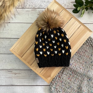 May include: Black knit beanie with a white and yellow heart pattern and a brown faux fur pom pom.