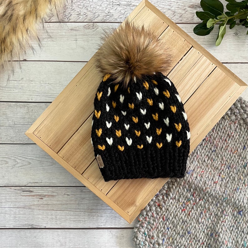 May include: Black knit beanie with a white and yellow heart pattern and a brown faux fur pom pom.