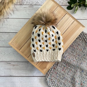May include: A white knitted beanie with a black and yellow heart pattern and a brown faux fur pom pom.
