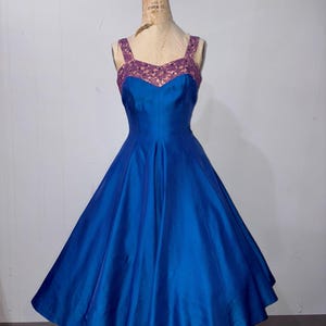 Puede incluir: Un vestido azul vintage con una falda amplia y un corpiño ajustado. El vestido presenta un escote corazón y tirantes anchos con detalles de encaje morado. El vestido se exhibe en un maniquí.