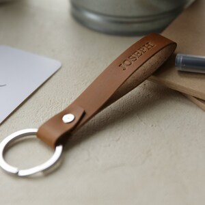 May include: A tan leather keychain with the name "JOSEPH" embossed on the strap. The keychain has a silver metal ring and a silver rivet. The background includes a pen, a card, and a metal container.