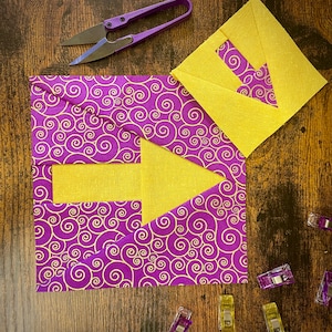 Op de afbeelding: Een paarse stof met een swirl-patroon is gelegd over een gele stof met een pijlvorm uitgesneden. De stof wordt bij elkaar gehouden met kleine clips.