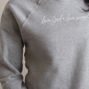A woman is tousling her hair while modeling a "love God + love people" grey sweatshirt