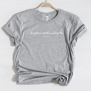 May include: Gray t-shirt with the text "His Grace Reaches Me" and the Bible verse "Romans 5:2" in white lettering.
