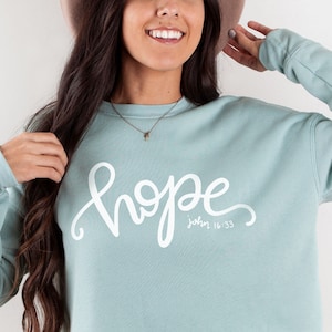 May include: A light blue sweatshirt with the word "hope" written in white cursive lettering. The text "John 16:33" is written in smaller white lettering below the word "hope".
