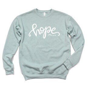 May include: A light blue sweatshirt with the word "hope" written in white cursive lettering. The text "John 14:33" is written in smaller white lettering below the word "hope".