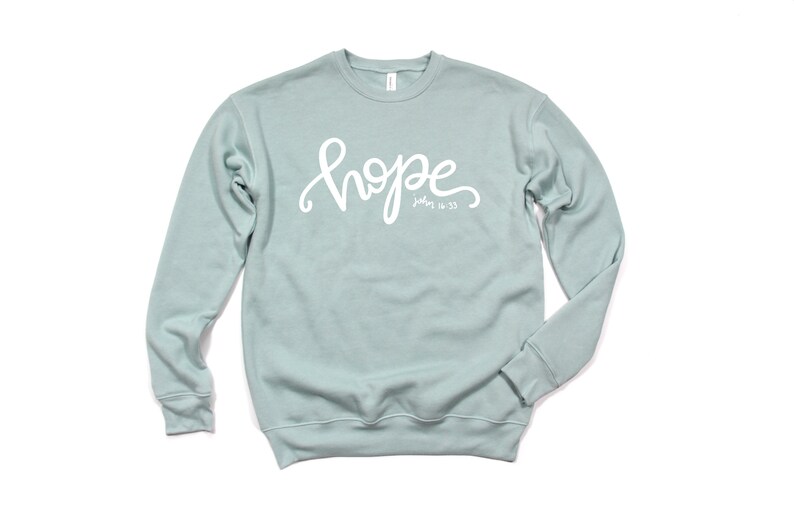 May include: A light blue sweatshirt with the word "hope" written in white cursive lettering. The text "John 14:33" is written in smaller white lettering below the word "hope".