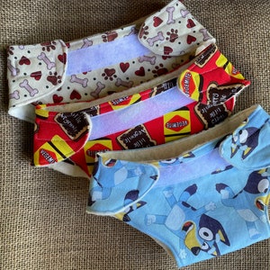 Puede incluir: Tres pañales reutilizables para perros con diferentes patrones. Uno es de color crema con huellas de patas y huesos marrones, otro es rojo con etiquetas de Vegemite, y el último es azul claro con personajes de dibujos animados de perros. Cada uno tiene un cierre de velcro blanco.