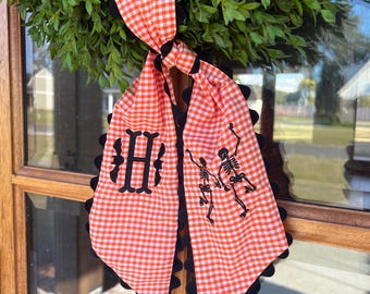 May include: Orange and white gingham scarf with black trim and embellishments. The scarf features a black monogram letter "H" and a dancing skeleton design, perfect for Halloween.