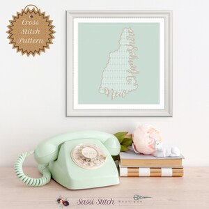 Op de afbeelding: Een ingelijst kruissteekpatroon van de staat New Hampshire met de woorden "I'd Rather Be In New Hampshire" geborduurd in lichtbruin garen op een lichtblauwe achtergrond.