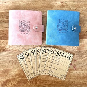 May include: Two faux leather seed storage notebooks, one pink and one blue, with the word "SEEDS" embossed on the front.  The notebooks are open to reveal blank seed tracking cards with the word "SEEDS" printed at the top.