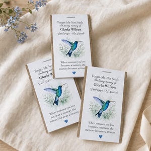 May include: Three rectangular seed packets featuring a watercolor hummingbird illustration, the text Forget-Me-Not Seeds, In loving memory of Gloria Wilson, 5/10/1940 - 8/14/2026, and the quote When someone you love becomes a memory, the memory becomes a treasure.