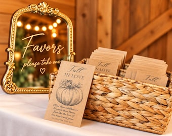 May include: A gold ornate mirror with the text Favors please take one and a heart icon, next to a woven basket filled with brown paper seed packets featuring a pumpkin illustration and the text Fall IN LOVE.