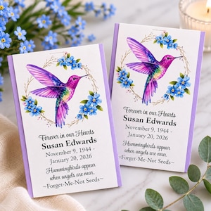 May include: Two rectangular memorial seed packets with light purple borders. Each features a vibrant purple and green hummingbird illustration surrounded by blue forget-me-not flowers. Text reads: Forever in our Hearts, Susan Edwards, November 9, 1944 - January 20, 2026, Hummingbirds appear when angels are near, Forget-Me-Not Seeds.