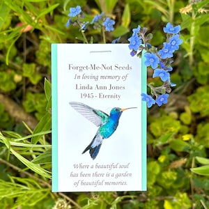 May include: A blue and green hummingbird with a long beak is printed on a light blue card with white text. The text reads "Forget-Me-Not Seeds In loving memory of Linda Ann Jones 1945 - Eternity Where a beautiful soul has been there is a garden of beautiful memories."