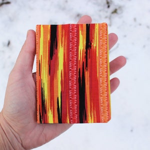 May include: A small, rectangular fabric notebook cover with a colourful abstract pattern in shades of red, orange, yellow, and black.